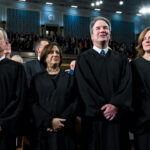 Four judges in black robes stand together in a large, ornate hall with an audience seated and standing behind them. The hall has blue patterned walls, bright lighting, and high ceilings.