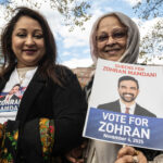 Two women outdoors hold a Vote for Zohran campaign sign and a phone. One wears a shirt with the candidates photo; both are smiling. There are trees and a blue sky in the background.