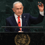 Benjamin Netanyahu in a suit and red tie stands at a United Nations podium, holding up a small black object with his right hand during a speech, with the UN emblem visible on the podium.
