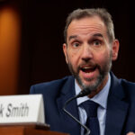 Jack Smith in a suit speaks into a microphone at a hearing. A nameplate in front of him reads Mr. Jack Smith. He has short hair and a beard and appears to be responding to a question.