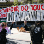 Two people hold a large banner reading Stanford, do you care about international law? with a painted Palestinian flag. Others stand in the background outside a Stanford campus building.
