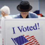 An elderly person wearing a black hat votes at a polling station this week, standing behind a booth labeled I Voted! with an American flag. Other voters are visible in the background.
