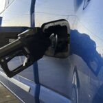 A black gas pump nozzle is inserted into the fuel tank of a blue vehicle at a gas station, with part of the truck and fuel door visible. The scene is lit by daylight.