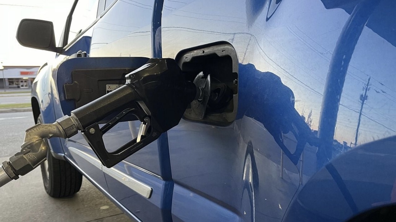A black gas pump nozzle is inserted into the fuel tank of a blue vehicle at a gas station, with part of the truck and fuel door visible. The scene is lit by daylight.