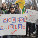 A group of people protest outdoors, holding signs with messages like SCHOOL CHOICE, STOP, and Danbury deserves a charter school. Some wear sunglasses and coats on a cloudy day.