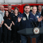 JD Vance speaks at a podium labeled Vice President of the United States while a group of people, some in uniforms, stand behind him in front of a red emergency vehicle.