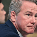 A man with short gray hair in a blue suit looks slightly to the side with a serious expression. Another person sits blurred in the background. An American flag is partially visible on the right.