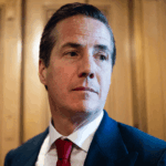 A man in a dark suit, white shirt, and red tie stands in front of wooden panels, looking slightly to his right with a neutral expression.