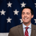Frank LaRose in a suit and red striped tie smiles in front of a backdrop with white stars on a blue background, resembling part of the American flag.