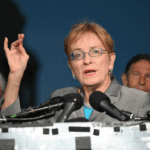 Marcy Kaptur in a gray suit and glasses speaks at a podium with microphones, gesturing with her hand. Several men in suits stand behind her. The background is dark, with a blurred building visible on the left.