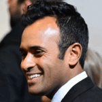 Vivek Ramaswamy in a black suit and white shirt smiles as he stands indoors, with blurred people and neutral-colored walls in the background.