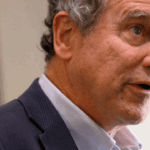 A man with gray hair wearing a dark blazer and light-colored shirt speaks, looking slightly to the side. The background is softly blurred, focusing attention on his face and expression.