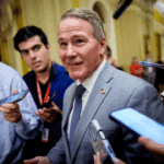 Jon Husted in a gray suit speaks to reporters about the upcoming midterms, as they surround him with phones and microphones in a hallway with yellow walls.