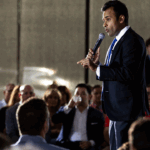 A man in a suit speaks into a microphone, addressing an attentive seated audience in a well-lit indoor setting.