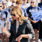 Amy Acton in sunglasses speaks into a microphone at an outdoor rally, surrounded by people holding Dr. Amy Acton for Governor signs. Many attendees wear blue shirts and sunglasses, showing their support.