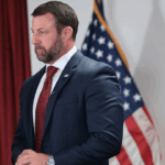 A man in a navy suit and red tie stands in front of a U.S. flag and a Department of Homeland Security seal, looking serious. A red curtain is visible to the left.
