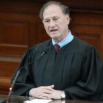 A judge in a black robe speaks at a podium in a courtroom, with a microphone in front of him and wooden paneling in the background.