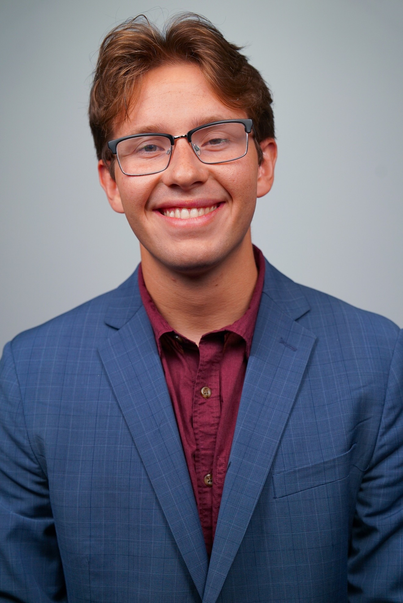 Daily Signal Internship 4 A young man with light brown hair, glasses, and a blue suit jacket over a maroon shirt—dressed for success in his daily signal internship—smiles at the camera against a plain light background.