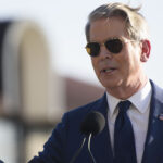 A man in a blue suit and sunglasses speaks at a podium outdoors, gesturing with one finger raised. An American flag is visible in the background.