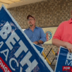 Two men hold Beth Macy for Congress signs outside a building, possibly following recent debates. One man wears a red shirt and beige hat, holding a small white dog. Another man in blue stands in the background, and a pet store logo is visible.