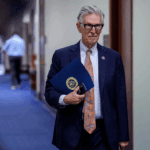 An older man in a suit and glasses holds a blue folder and sunglasses, walking down a hallway with blue carpet and wooden doors—perhaps on his way to discuss the latest developments in AI.