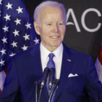 Joe Biden in a blue suit speaks at a podium with microphones, standing between two American flags, with a black background featuring partial white text.