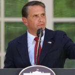 A man in a dark suit and red tie speaks at a podium outdoors, gesturing with his right hand as he addresses concerns about election fraud. American flags and green foliage are in the background.