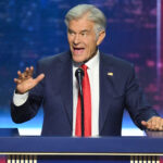 Dr. Mehmet Oz in a suit and red tie speaks energetically at a podium, gesturing with his left hand. The background is blue with white stars and red stripes, resembling a patriotic or political setting.