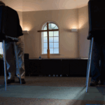 Two people stand at separate voting booths in a room with carpeted floors, sunlight streaming in, and a cross and windows visible in the background.