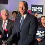 Senator John Thune is pictured in a suit and speaks in front of reporters' microphones. Several people stand behind him holding Mike Collins U.S. Senate signs. The setting is indoors with red lamps.