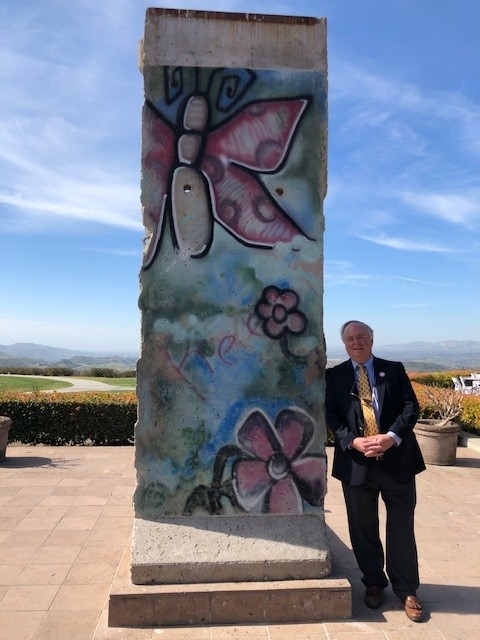 Reagan’s Berlin Wall Speech Forever Changed the World 1 A man in a suit stands next to a tall, decorated cement slab featuring butterfly and flower graffiti—an artwork reminiscent of the Reagan era—with hills and blue sky in the background.