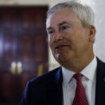A man in a suit and red striped tie stands indoors, smiling slightly, with a blurred wooden door and a cream-colored wall in the background.