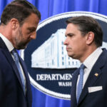 Two men in suits face each other seriously in front of a Department of Justice sign, with an American flag in the background.