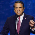 Todd Blanche in a dark suit and red tie speaks passionately at a microphone, gesturing with his right hand, against a blurred blue background.