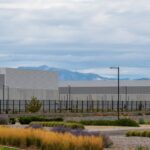 A large, modern industrial data center building with gray walls and flat roofs. It sits behind a black fence, surrounded by landscaping with grasses and shrubs, with mountains and cloudy skies in the background.
