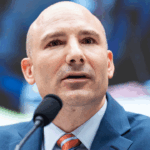 Steve Descano speaks into a microphone at a formal event, with blurred people in the background.