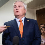 James Comer in a blue suit and orange tie stands in a hallway, surrounded by people and microphones, as reporters hold out recording devices and take notes.