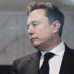 Elon Musk in a black suit and tie looks to the side with a neutral expression while standing indoors.