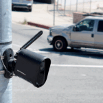 A black security camera is mounted on a metal pole beside a road, with a gray pickup truck driving by in the background.