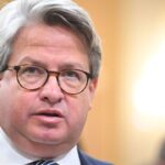 Gabe Sterling, Director of Special Projects for Secretary of State Brad Raffensperger, appears with glasses, wearing a dark suit and tie. He speaks indoors with a blurred background and bright lighting.
