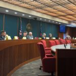 Members of the Joint Election Equipment Specifications and Standards Committee sit at a long, curved table at their third meeting with red chairs, U.S. and state flags, microphones, and nameplates.