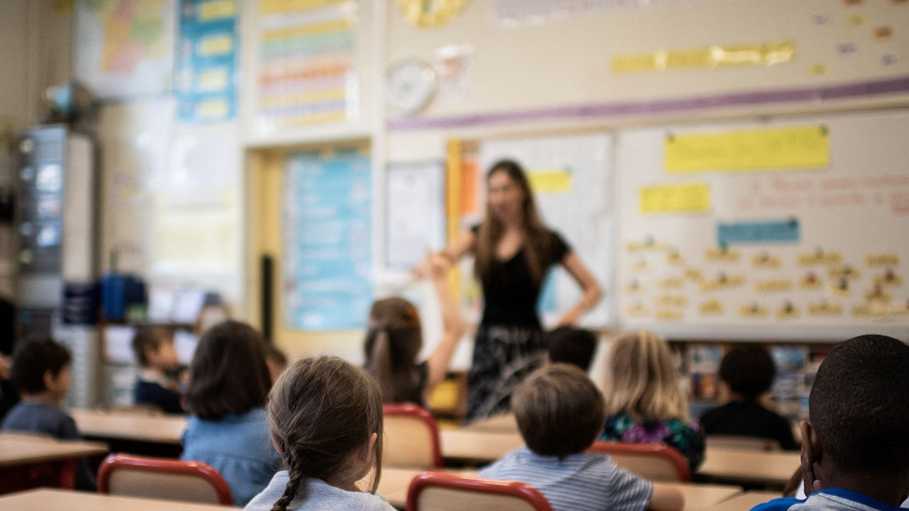 A classroom with young students seated at desks, facing a teacher standing at the front near a whiteboard covered in colorful posters and notes, while a discussion about violent books sparks thoughtful conversation among the class.
