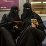 Two people wearing black niqabs sit on a subway bench in America. One holds a phone, and the other holds a cup with a straw. Subway poles and seats are visible, along with a purple ad in the background.