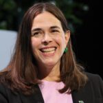 A woman with long brown hair, wearing a black blazer and pink top, smiles while seated indoors. Green plants are visible in the background.