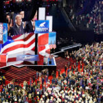 A large crowd fills an arena with a stage displaying RNC 2024 and American flags. A speaker addresses the audience, while people hold signs and cheer. The atmosphere is lively and patriotic.
