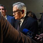 A man in a suit and glasses stands surrounded by reporters and microphones, facing a group of people in a dimly lit indoor setting.