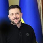 A man with short dark hair and a trimmed beard smiles while standing in front of blurred blue and yellow flags. He wears a black shirt with an emblem on the left side.