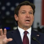 A man in a dark suit and red tie gestures while speaking, with an American flag in the blurred background.