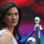 A woman with long dark hair wearing a light blue dress and a red lanyard looks toward the camera, with a blurred background of blue and red hues.