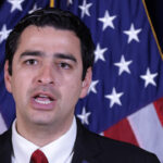 Gabe Evans in a suit and red tie speaks in front of two American flags, appearing to address an audience or the media.
